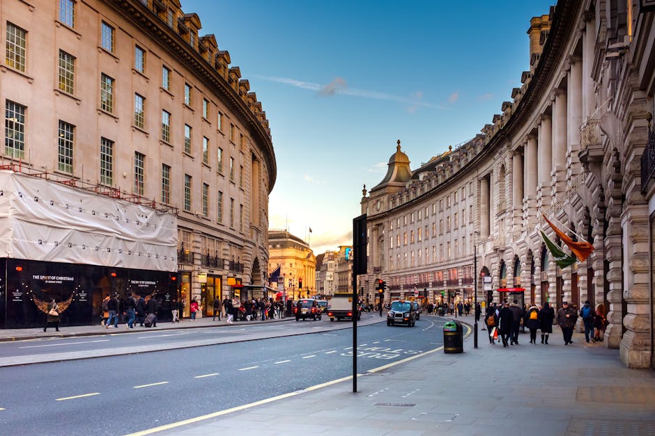 A busy urban street scene in Central London featuring a mix of historic and modern buildings. On the right, a white stone building with ornate window frames and decorative details contrasts with a nei