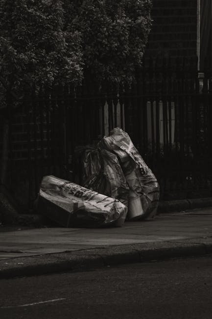 A narrow urban alleyway filled with a large black and beige wheeled garbage bin positioned in the foreground, surrounded by scattered waste and debris on the ground. Behind the bin, there are overflow