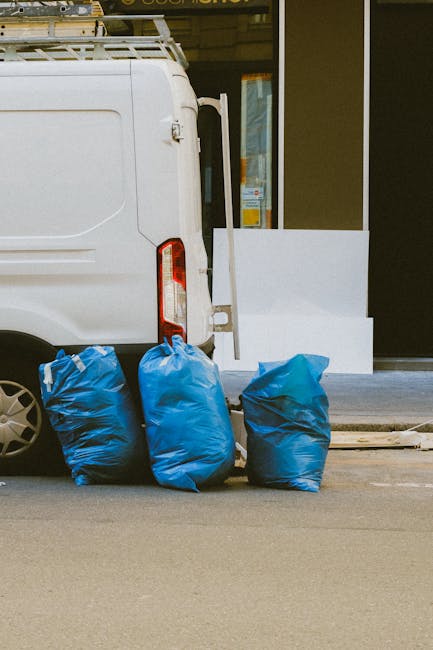 A man wearing a grey beanie, black jacket, and high-visibility yellow vest stands beside a large metal trolley loaded with various types of rubbish, including black, grey, and orange trash bags and as